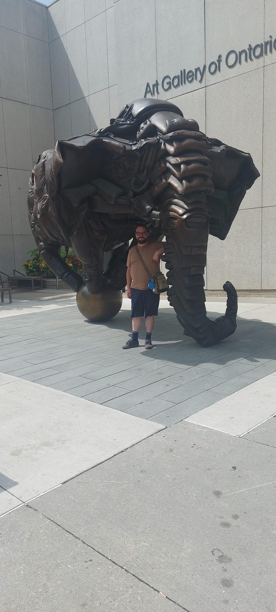 A man short hair and bushy facial hair leans on the trunk of a brown elephant made of leather couch cushons standing on a ball